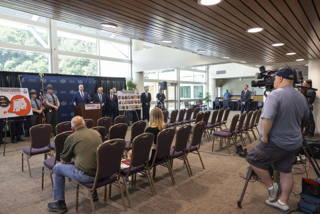 everal law enforcement officials and other personnel stand behind the podium, flanked by two display boards labeled “Operation Summer Street Sweeper,” one showing a map and the other showing multiple headshot photos.