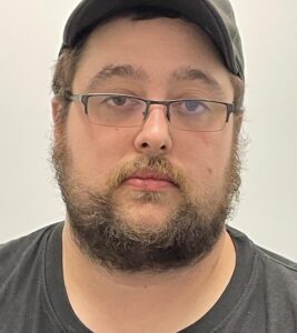 A portrait-style photograph of an individual facing the camera against a plain light-colored background. The individual is wearing glasses, a dark shirt, and a dark cap, and has short facial hair.
